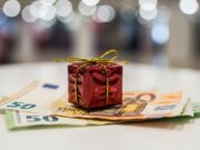 Regali di Natale last minute: soluzioni utili sotto i 50 euro per non sbagliare Primo piano drammatico di un piccolo regalo di Natale su banconote, simbolo di acquisti last minute sotto i 50€.