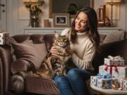 I 10 migliori regali per gli amanti dei gatti che hanno già tutto Una donna sorride mentre il suo gatto di razza Maine Coon dorme su un divano di lusso, con regali a tema gatto sullo sfondo.