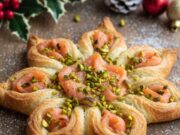 Stella di sfoglia salmone e pistacchio: l’antipasto di Natale pronto in 15 minuti che fa fare un figurone Elegante stella di sfoglia salata ripiena di salmone affumicato e decorata con granella di pistacchio su un piatto natalizio.