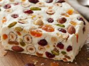 Torrone senza cottura: il regalo dell’ultimo minuto pronto in freezer Fetta di torrone senza cottura, cremoso e ripieno di frutta secca, presentato su un tagliere di legno rustico.