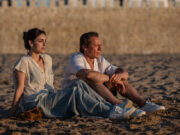 Le cose non dette: un ultimo bacio per Muccino? Miriam Leone e Stefano Accorsi seduti sulla sabbia di una spiaggia al tramonto, con un muro di pietra antica sullo sfondo. Entrambi guardano verso l'orizzonte in un momento di riflessione.