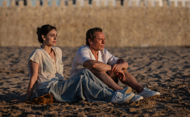 Miriam Leone e Stefano Accorsi seduti sulla sabbia di una spiaggia al tramonto, con un muro di pietra antica sullo sfondo. Entrambi guardano verso l'orizzonte in un momento di riflessione.
