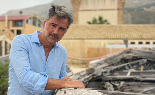 Beppe Convertini in posa appoggiato a un'antica imbarcazione in legno con sfondo architettonico.