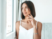 I gioielli in argento da donna tornano protagonisti: quali sono i must have Donna con top bianco che indossa un bracciale rigido in argento vicino a una finestra