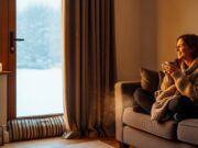 Bollette leggere: 3 trucchi “invisibili” per isolare casa a costo zero Donna sorridente su un divano in un salotto caldo con tenda pesante e paraspifferi alla porta finestra in inverno.