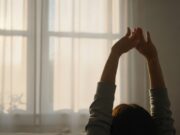 Svegliarsi presto: perché seguire il proprio ritmo biologico è cruciale Primo piano ultra-realistico di una persona serena che si allunga, risvegliata naturalmente dal proprio ritmo biologico. La luce dorata e diffusa del sole di metà mattina (circa le 10:00) filtra attraverso le tende di lino, creando un'atmosfera di calma e riposo ottimale.