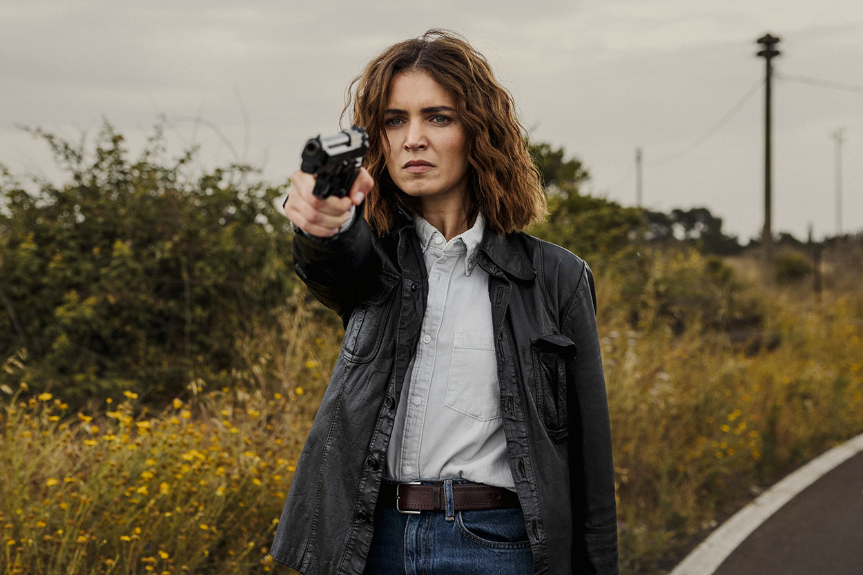 Giusy Buscemi, nel ruolo della vicequestore Vanina Guarrasi, indossa una giacca di pelle nera e punta una pistola in avanti con espressione decisa, inquadrata su una strada di campagna sotto un cielo nuvoloso.