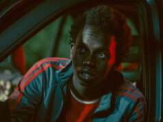 Cattiva strada: l’esordio di Davide Angiuli tra la criminalità barese e i legami di sangue Ritratto cinematografico di Malich Cissé all'interno di un'auto di notte, illuminato da contrastanti luci soffuse rosse e verdi. Indossa una giacca sportiva verde con strisce rosse e ha un'espressione intensa, mentre guarda fuori dall'inquadratura con gli occhi spalancati.