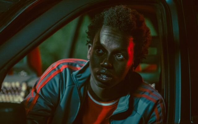 Ritratto cinematografico di Malich Cissé all'interno di un'auto di notte, illuminato da contrastanti luci soffuse rosse e verdi. Indossa una giacca sportiva verde con strisce rosse e ha un'espressione intensa, mentre guarda fuori dall'inquadratura con gli occhi spalancati.