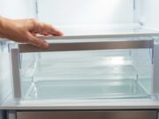 Acqua sotto i cassetti del frigorifero: come liberare il foro di scolo e salvare il compressore Pozzanghera d'acqua trasparente sotto il cassetto della verdura di un frigorifero aperto da una mano.