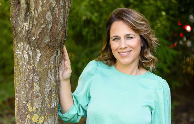 Ritratto all'aperto dell'attrice e conduttrice Annamaria Fittipaldi: sorridente e in posa rilassata con una mano appoggiata al tronco di un albero, indossa una blusa verde acqua su uno sfondo naturale sfocato
