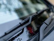 Liquido tergicristalli fai-da-te: perché l’acqua del rubinetto brucia la pompa lavavetri Dettaglio di spazzole tergicristalli e gocce d'acqua sul parabrezza di un'auto scura.