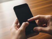 Telefono bloccato sulla scritta iniziale: come fare il riavvio di emergenza senza perdere i dati Mani di una persona che premono i tasti del volume e di accensione su uno smartphone spento appoggiato su una scrivania di legno.