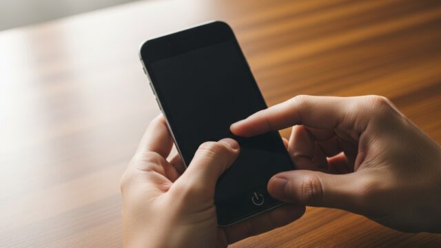 Mani di una persona che premono i tasti del volume e di accensione su uno smartphone spento appoggiato su una scrivania di legno.