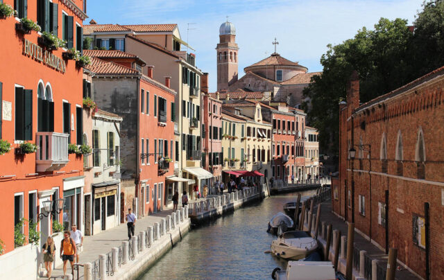 Ecco il tag alt perfetto per l'immagine: ottimizzato, descrittivo e della lunghezza ideale per l'accessibilità e la SEO. Tag Alt: "Veduta di un canale tranquillo a Venezia con persone che camminano lungo la fondamenta, edifici storici colorati e un campanile sullo sfondo.