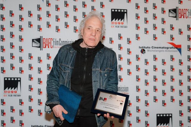 Il regista Abel Ferrara posa con il Premio Dino Ceccuzzi all'eccellenza cinematografica davanti al backdrop ufficiale del B.A. Film Festival.