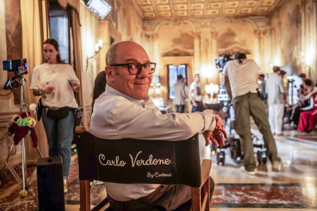 Carlo Verdone sul set del film "Scuola di seduzione" seduto sulla sedia da regista personalizzata in un interno d'epoca.