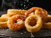 Frittura di pesce unta e molliccia: gli errori di fisica e chimica che distruggono la croccantezza Calamari e gamberi fritti dorati e croccanti appoggiati su una griglia di scolo in metallo, illuminazione drammatica laterale.
