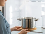 Sbrinare il freezer: perché usare il phon o il coltello distrugge irreparabilmente l’elettrodomestico Donna che inserisce una pentola d'acciaio fumante su un sottopentola in sughero all'interno di un frigorifero aperto per sbrinare il freezer con il metodo del vapore.