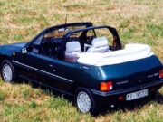 Peugeot 205 cabriolet: storia, quotazioni e vantaggi auto d’epoca Vista posteriore di tre quarti di una Peugeot 205 Cabriolet storica verde scuro, parcheggiata su un prato d'erba secca. L'auto è decappottabile, con la capote in tela ripiegata bianca