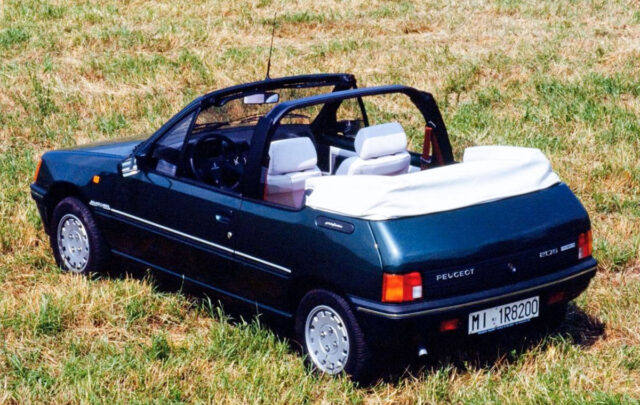Vista posteriore di tre quarti di una Peugeot 205 Cabriolet storica verde scuro, parcheggiata su un prato d'erba secca. L'auto è decappottabile, con la capote in tela ripiegata bianca