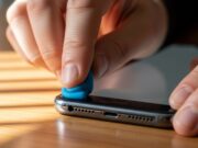 Audio dello smartphone basso: l’errore dello stuzzicadenti che distrugge il telefono Un primo piano macro dettagliato di un dito di una persona anziana che preme con precisione un pezzo di gomma adesiva blu sopra i fori dell'altoparlante inferiore di uno smartphone scuro, su una scrivania di legno.