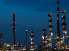 Riqualificazione energetica per le aziende: cosa c’è da sapere Veduta notturna di un complesso industriale con grandi ciminiere a strisce bianche e rosse illuminate sotto un cielo blu profondo.