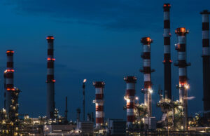 Riqualificazione energetica per le aziende: cosa c’è da sapere Veduta notturna di un complesso industriale con grandi ciminiere a strisce bianche e rosse illuminate sotto un cielo blu profondo.