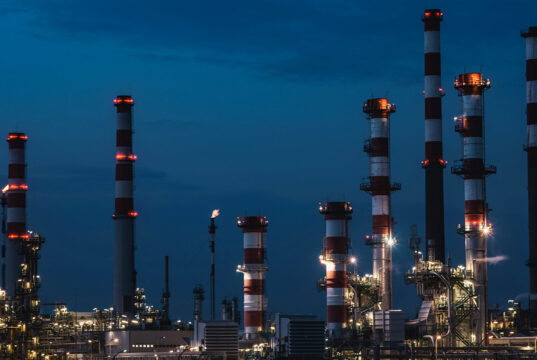 Riqualificazione energetica per le aziende: cosa c’è da sapere Veduta notturna di un complesso industriale con grandi ciminiere a strisce bianche e rosse illuminate sotto un cielo blu profondo.