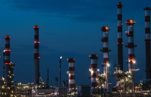 Veduta notturna di un complesso industriale con grandi ciminiere a strisce bianche e rosse illuminate sotto un cielo blu profondo.