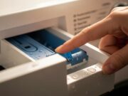 Il tasto segreto della lavatrice: a cosa serve la linguetta nel cassetto Primo piano di una mano che preme la linguetta azzurra per sbloccare il cassetto dei detersivi di una lavatrice.