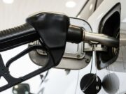 L’abitudine di forzare il pieno di benzina dopo il click che distrugge il filtro canister Dettaglio macro della pistola erogatrice nera inserita nel bocchettone di un'auto grigia, con benzina liquida che tracima e cola lungo la carrozzeria sotto il tappo appeso bagnato.