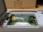 Giochi vintage in soffitta: l’errore delle pile che sta distruggendo il tuo tesoro anni ’90 Inquadratura macro autentica della corrosione bianca e verde causata dalle vecchie pile AA nel vano batterie di un gioco elettronico portatile vintage