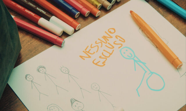 Un foglio bianco su un tavolo di legno circondato da pennarelli colorati, con la scritta arancione "NESSUNO ESCLUSO" e disegni stilizzati di figure umane a pennarello, tra cui una figura sorridente in azzurro che spicca rispetto alle altre.