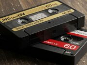Videocamere a cassetta MiniDV: come usarle oggi e l’errore che distrugge i tuoi vecchi nastri Macro iper-realistica di due cassette MiniDV (JVC nera e rossa) impolverate su tavolo di legno con muffa visibile sul nastro magnetico.