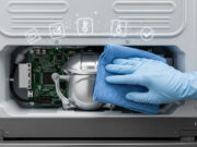 Frigorifero smart bloccato o che non raffredda: come fare l’hard reset Mano con guanto blu che pulisce con un panno il compressore e la scheda madre sul retro di un frigorifero smart per risolvere un blocco del sistema, con icone diagnostiche in sovrimpressione.
