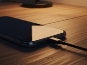 L’errore della ricarica notturna che distrugge la batteria dello smartphone Primo piano di uno smartphone moderno appoggiato su un tavolo in legno scuro mentre viene ricaricato tramite un cavo USB-C nero in un ambiente domestico con luce calda e soffusa.