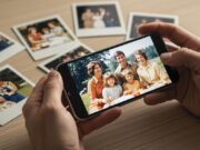 Google Foto AI Enhance distrugge le foto d’epoca: come proteggere i ricordi di famiglia Mani di una persona che tiene uno smartphone con una vecchia foto di famiglia modificata dall'intelligenza artificiale, appoggiato su una scrivania con polaroid originali.