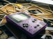 Nostalgia anni ’90: perché non riesci a smettere di tornarci e cosa dice di te Vecchio Game Boy viola trasparente impolverato su una scrivania disordinata tra cavi e fotografie vintage.