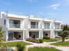 Piastrelle 30×120: il formato che valorizza gli spazi contemporanei Un complesso residenziale moderno di colore bianco con ampie balconate in vetro, giardini ben curati e un ulivo centenario, sotto un cielo azzurro e soleggiato.