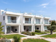 Piastrelle 30×120: il formato che valorizza gli spazi contemporanei Un complesso residenziale moderno di colore bianco con ampie balconate in vetro, giardini ben curati e un ulivo centenario, sotto un cielo azzurro e soleggiato.