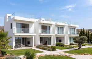Piastrelle 30×120: il formato che valorizza gli spazi contemporanei Un complesso residenziale moderno di colore bianco con ampie balconate in vetro, giardini ben curati e un ulivo centenario, sotto un cielo azzurro e soleggiato.