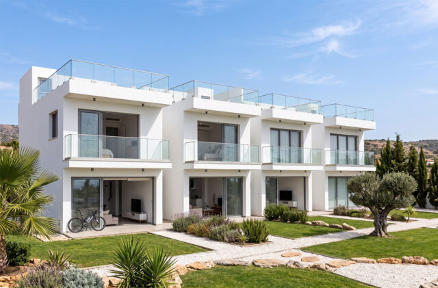 Un complesso residenziale moderno di colore bianco con ampie balconate in vetro, giardini ben curati e un ulivo centenario, sotto un cielo azzurro e soleggiato.