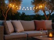 Il salotto sotto le stelle: idee per arredare la zona relax serale in giardino Divano da giardino con cuscini in lino e terracotta, calice di vino e candela su tavolino in legno, luci festoon tra gli ulivi al tramonto