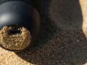 Cassa Bluetooth in spiaggia: l’errore con sabbia e caldo che la rovina in una stagione Dettaglio macro di una cassa bluetooth nera con la porta usb-c ostruita da granelli di sabbia sotto il sole estivo.