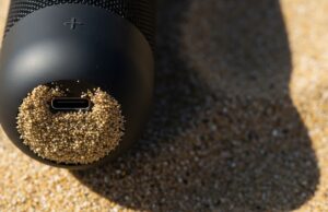 Cassa Bluetooth in spiaggia: l’errore con sabbia e caldo che la rovina in una stagione Dettaglio macro di una cassa bluetooth nera con la porta usb-c ostruita da granelli di sabbia sotto il sole estivo.