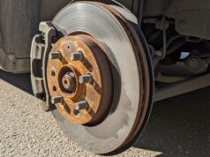 Auto in vacanza per settimane: perché il freno a mano tirato può incollare le pasticche ai dischi Disco freno arrugginito con ossidazione arancione sul mozzo, ripreso dal basso su asfalto con luce solare diretta, auto ferma in estate