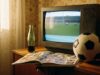Ciao, la mascotte di Italia ’90: il dettaglio che (quasi) nessuno nota Vecchia TV a tubo catodico anni '90 che trasmette una partita di calcio, con album di figurine, pallone e bottiglia di vetro in un salotto d'epoca