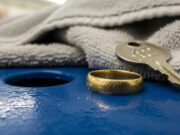 Nuoti con la fede al dito? Il cloro rovina l’oro in un modo che non vedi subito Fede nuziale d'oro graffiata appoggiata su una superficie bagnata accanto alla chiave dell'armadietto in piscina