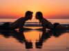 Perché in vacanza ci si innamora più facilmente (e come capire se è reciproco) Coppia sdraiata sulla spiaggia al tramonto, sguardi speculari che comunicano un'attrazione reciproca durante le vacanze estive